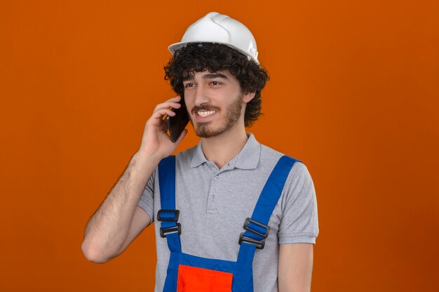 격리 된 오렌지 벽을 통해 행복 한 얼굴로 웃 고 휴대 전화에 얘기하는 건설 유니폼과 안전 헬멧을 착용하는 젊은 수염 잘 생긴 작성기