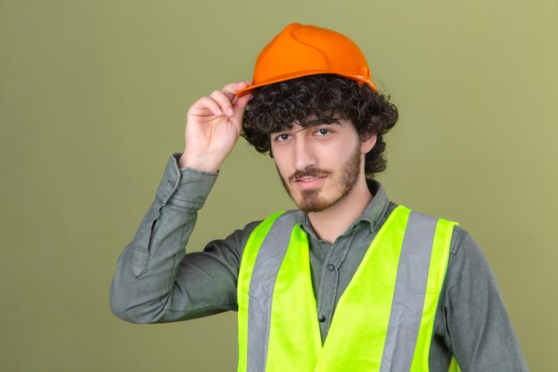 Young bearded handsome engineer making greeting gesture touching his helmet smiling standing over isolated green wall