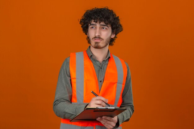 Молодой бородатый красивый инженер в строительном жилете держит в руках буфер обмена, пишет что-то, глядя в сторону с задумчивым выражением лица над изолированной оранжевой стеной