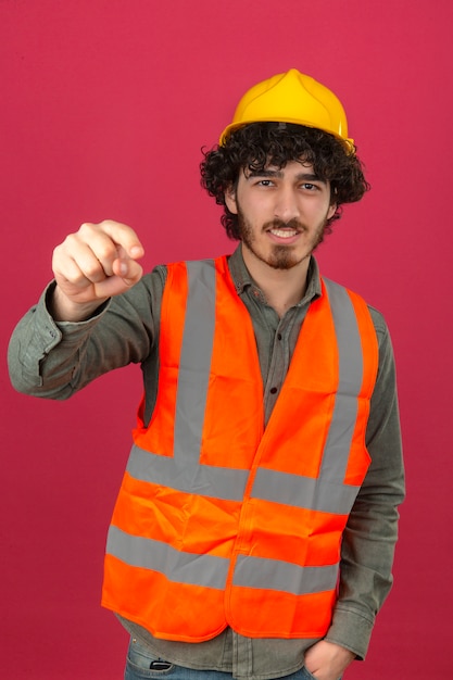 Young bearded handsome engineer wearing security helmet and vest pointing displeased and frustrated to the camera angry and furious with you over isolated pink wall