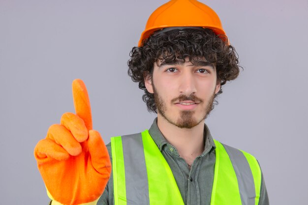 Young bearded handsome engineer wearing security helmet and vest showing number one sign with smile over isolated white wall
