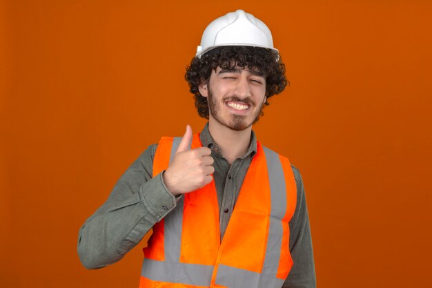 Молодой бородатый красивый инженер в шлеме безопасности и жилете, весело улыбаясь, подмигивая, показывая большой палец вверх над изолированной оранжевой стеной