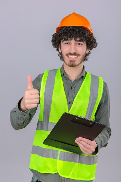 Young bearded handsome engineer wearing security helmet and vest with smile on face showing thumb up over isolated white wall