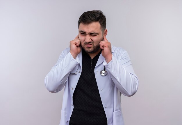 Young bearded male doctor wearing white coat with stethoscope closing ears with fingers  with annoyed expression for the noise of loud sound 