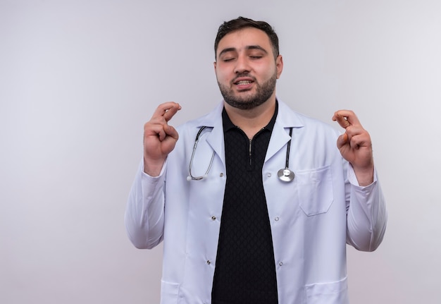 Young bearded male doctor wearing white coat with stethoscope closing eyes making desirable with crosing fingers 