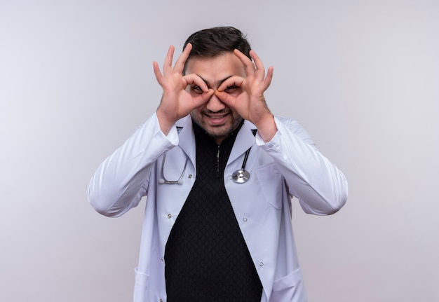 Young bearded male doctor wearing white coat with stethoscope doing ok signs  with fingers as binoculars looking through fingers smiling having fun 