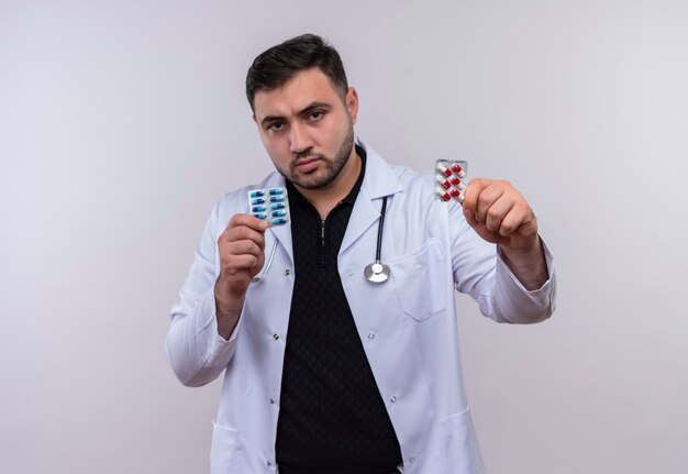 Young bearded male doctor wearing white coat with stethoscope holding blisters with pills showing to camera with serious face 