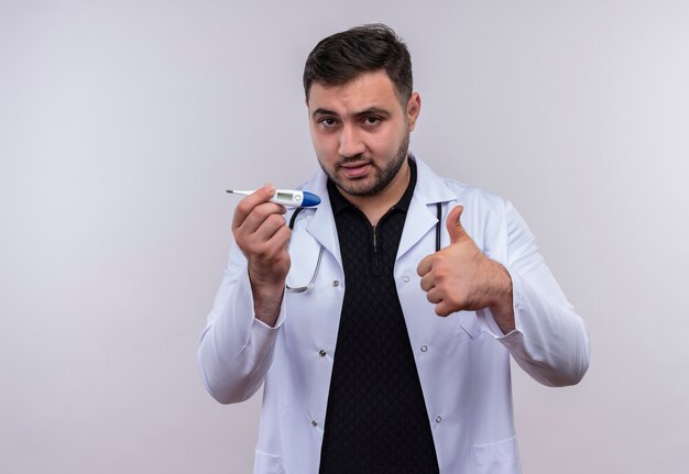 Молодой бородатый мужчина-врач в белом халате со стетоскопом держит цифровой термометр, уверенно улыбаясь, показывает палец вверх