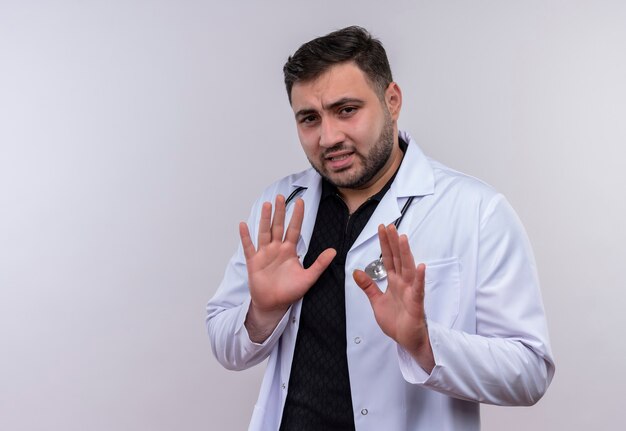 Молодой бородатый мужчина-врач в белом халате со стетоскопом протягивает руки, делая защитный жест с выражением отвращения