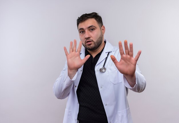 Young bearded male doctor wearing white coat with stethoscope holding out hands making defense gesture with fear expression 