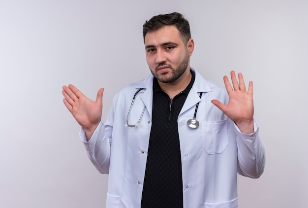 Young bearded male doctor wearing white coat with stethoscope holding out hands making defense gesture with fear expression 