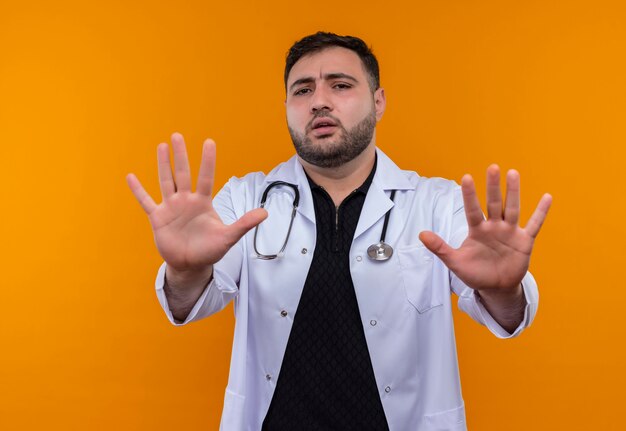 Young bearded male doctor wearing white coat with stethoscope holding out his hands making stop sign with fear expression 