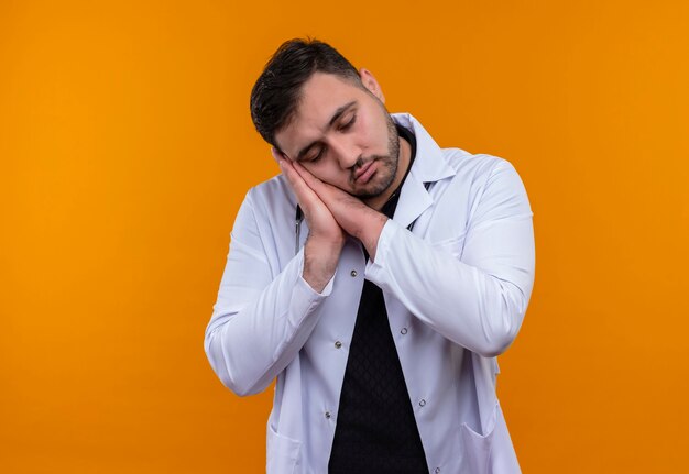 Giovane maschio barbuto medico indossa camice bianco con lo stetoscopio tenendo le palme insieme la testa pendente sui palmi vuole dormire