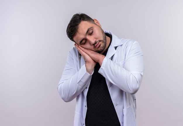 Young bearded male doctor wearing white coat with stethoscope holding palms together leaning head on palms wants to sleep