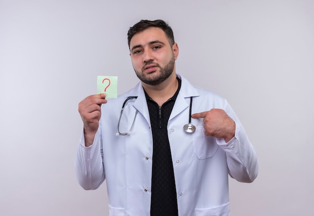 Young bearded male doctor wearing white coat with stethoscope holding reminder paper with question mark pointing with index finger to it 