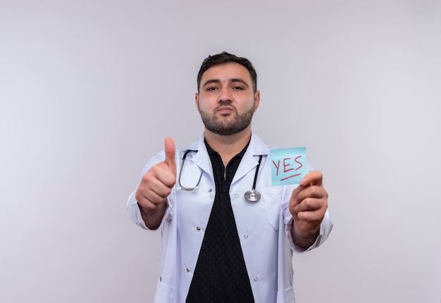 Young bearded male doctor wearing white coat with stethoscope holding reminder paper with yes word showing thumbs up 