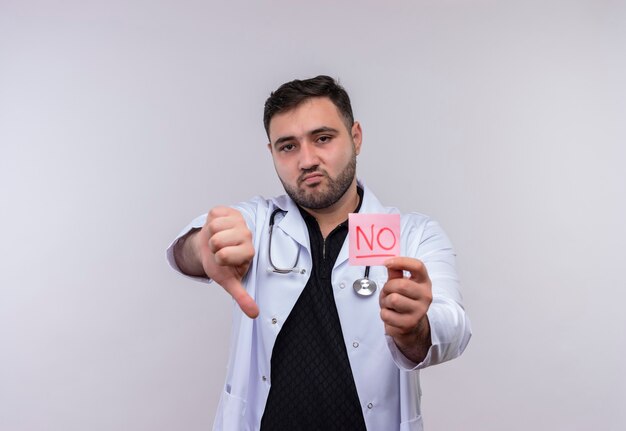 Young bearded male doctor wearing white coat with stethoscope holding remonder paper with word no looking displeased showing dislike 