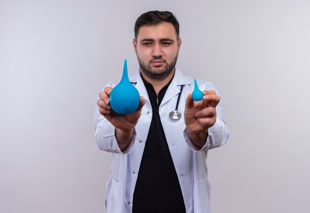 Young bearded male doctor wearing white coat with stethoscope holding small and large enemas showing to camera 