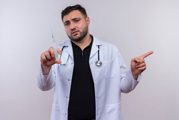 Молодой бородатый мужчина-врач в белом халате со стетоскопом держит шприц в замешательстве, указывая пальцем в сторону