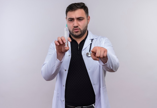 Young bearded male doctor wearing white coat with stethoscope holding syringe pointing with finger to camera with serious face 