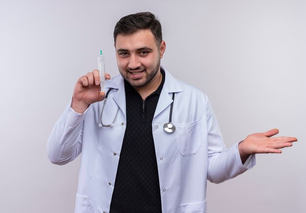 Foto gratuita giovane maschio barbuto medico indossa camice bianco con lo stetoscopio tenendo la siringa sorridente fiducioso