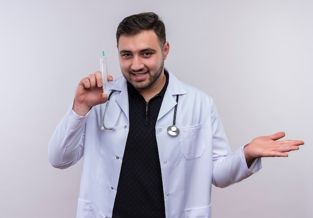 Young bearded male doctor wearing white coat with stethoscope holding syringe smiling confident 