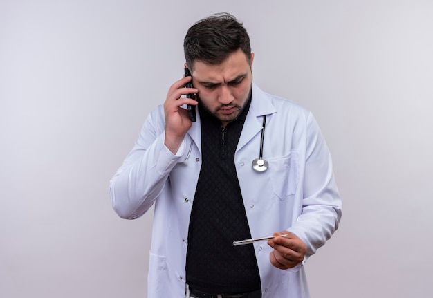 Young bearded male doctor wearing white coat with stethoscope holding thermometer  looking at it very emotional and worried while talking on mobile phone 