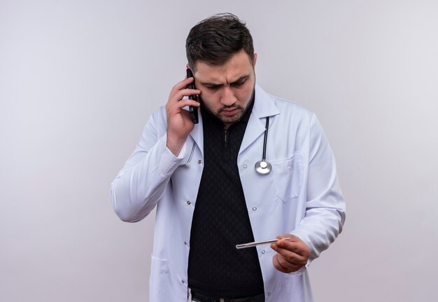 Молодой бородатый мужчина-врач в белом халате со стетоскопом, держащим термометр, смотрит на него очень эмоционально и обеспокоен во время разговора по мобильному телефону