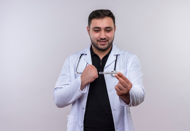 Free photo young bearded male doctor wearing white coat with stethoscope holding thermometer  looking at it with happy face