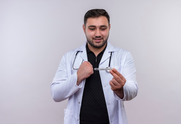 Молодой бородатый мужчина-врач в белом халате со стетоскопом держит термометр и смотрит на него с счастливым лицом