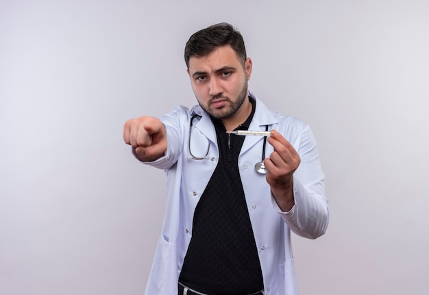 Молодой бородатый мужчина-врач в белом халате со стетоскопом держит термометр, указывая пальцем на камеру с серьезным лицом