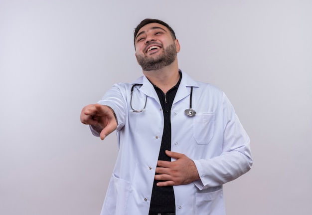 Молодой бородатый мужчина-врач в белом халате со стетоскопом шутит, смеется, высмеивает кого-то