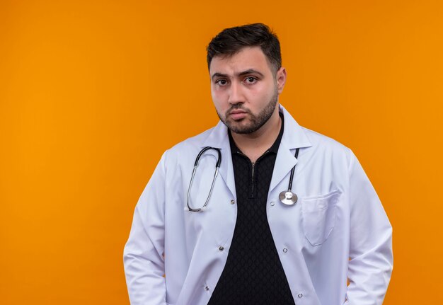 Young bearded male doctor wearing white coat with stethoscope loking at camera with serious face 