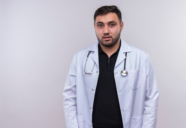 Free photo young bearded male doctor wearing white coat with stethoscope looking at camera confused