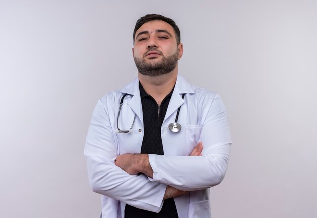 Young bearded male doctor wearing white coat with stethoscope looking at camera self-satisfied with arms crossed 