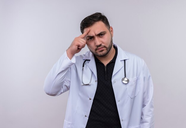 Молодой бородатый мужчина-врач в белом халате со стетоскопом смотрит в камеру с серьезным лицом недоволен