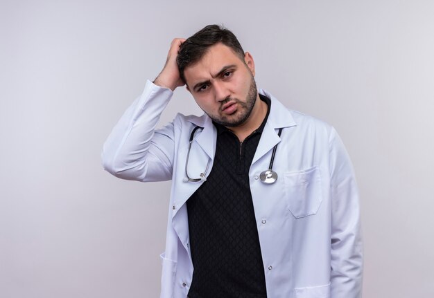 Молодой бородатый мужчина-врач в белом халате со стетоскопом выглядит смущенным, трогая голову рукой