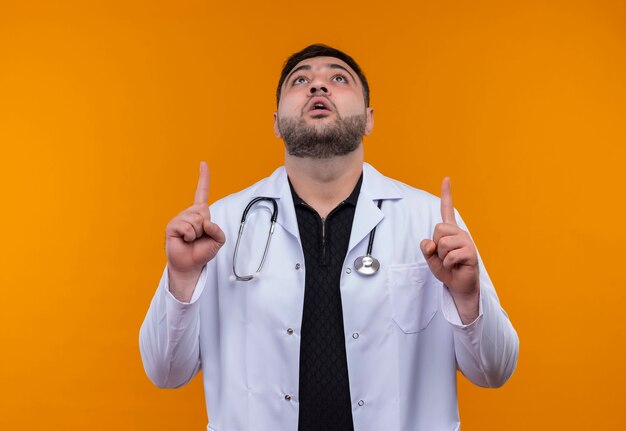 Young bearded male doctor wearing white coat with stethoscope looking surprised pointing with index fingers up 