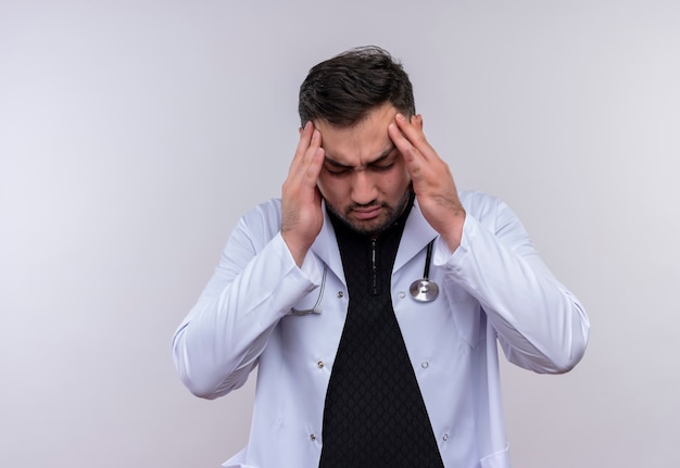 Молодой бородатый мужчина-врач в белом халате со стетоскопом выглядит усталым и скучающим с головной болью