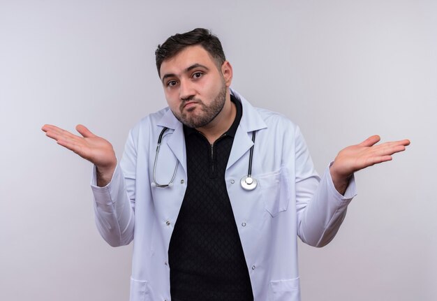 Young bearded male doctor wearing white coat with stethoscope looking uncertain and confused spreading arms to the sides 