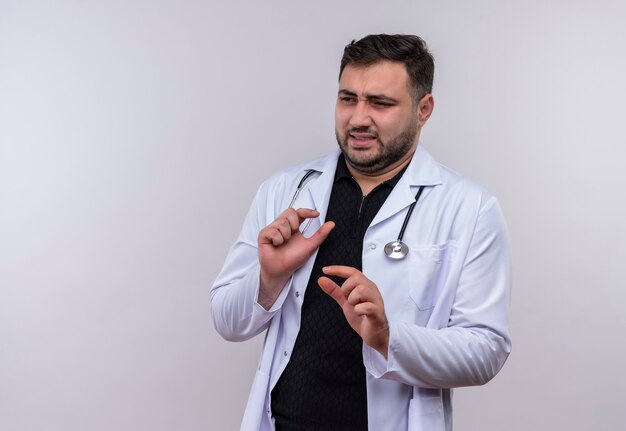 Young bearded male doctor wearing white coat with stethoscope making defense gesture with arms with dusgusted expression 