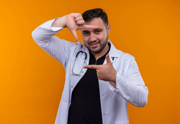 Young bearded male doctor wearing white coat with stethoscope making frame with fingers looking through this frame smiling 