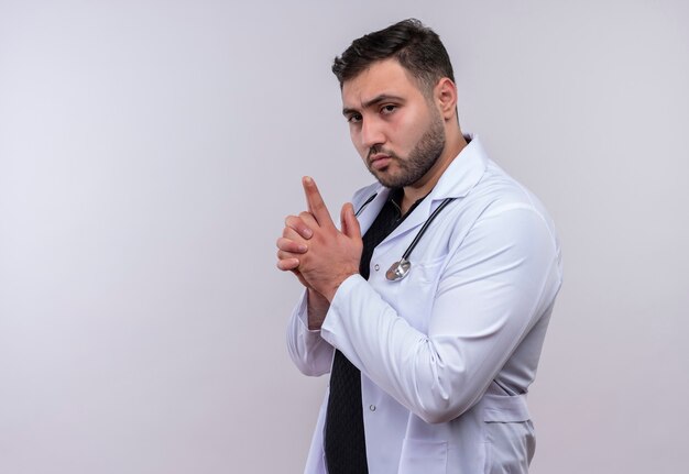 Young bearded male doctor wearing white coat with stethoscope making pistol or gun gesture with fingers looking confident 