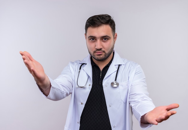Молодой бородатый мужчина-врач в белом халате со стетоскопом делает приветственный жест