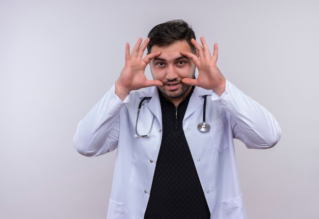 Молодой бородатый мужчина-врач в белом халате со стетоскопом, закрывая глаза пальцами