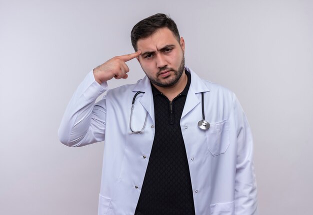 Young bearded male doctor wearing white coat with stethoscope pointing his temple looking at camera with saerious face focused on task 