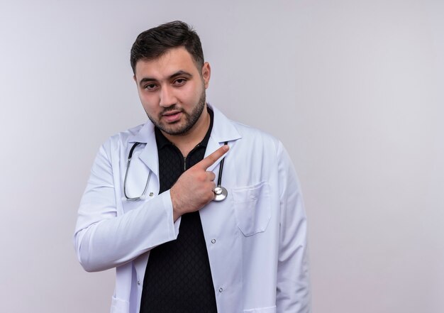 Young bearded male doctor wearing white coat with stethoscope pointing with finger to the side looking confident 