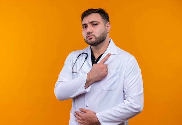 Молодой бородатый мужчина-врач в белом халате со стетоскопом, указывая указательным пальцем в сторону, выглядит уверенно