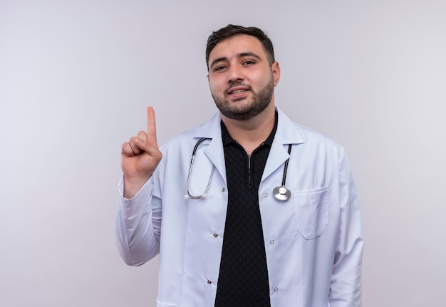 Молодой бородатый мужчина-врач в белом халате со стетоскопом, указывая указательным пальцем вверх, глядя в камеру с серьезным уверенным выражением лица