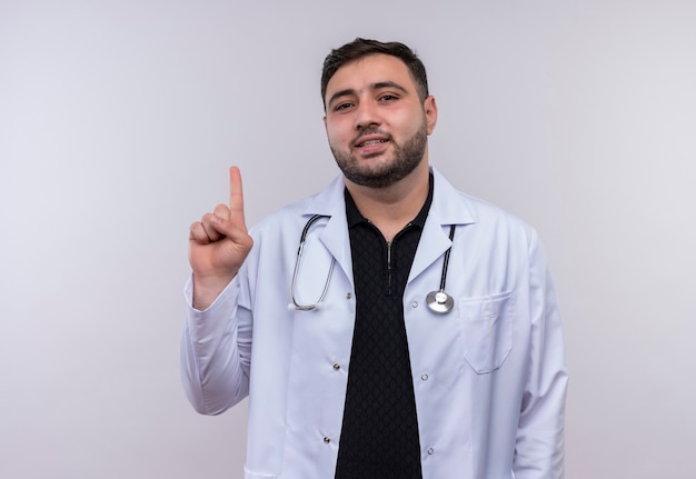 Young bearded male doctor wearing white coat with stethoscope pointing with index finger up  looking at camera  with serious confident expression 